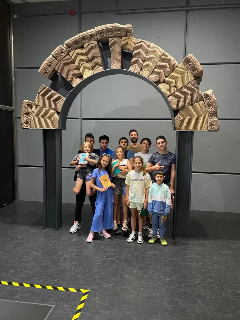 Grupo de niños y adultos posando frente a un arco decorativo en un espacio interior, mostrando sonrisas y sosteniendo cuadernos y materiales. El ambiente sugiere una actividad educativa o recreativa.
